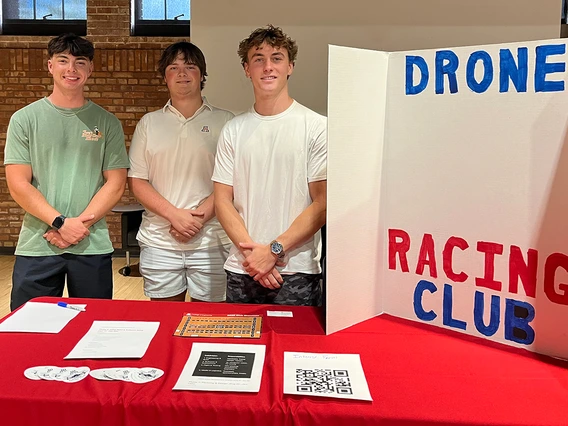 Three young men stand behind a red table with a poster board that reads: Drone Racing Club.