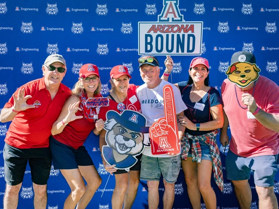 Six adults in multicolored shirts stand in front of a U of A College of Engineering photo wall.
