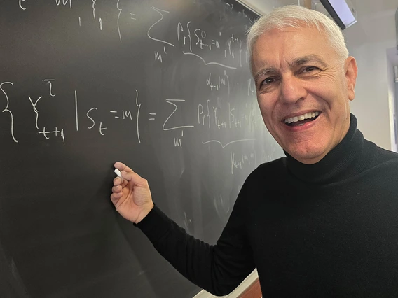 A man in a black turtleneck sweater stands in front of a black chalkboard.