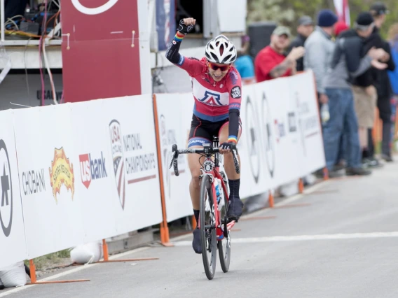 Erica Clevenger, on her bicycle wearing a UA jersey, raises her fist above her head in victory