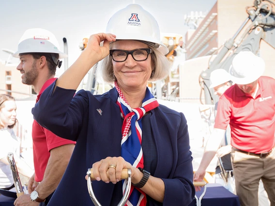 Betsy Cntwell on a construction site, wearing a white hard hat with a Block A logo. She is smiling and holding the bill of the hat.