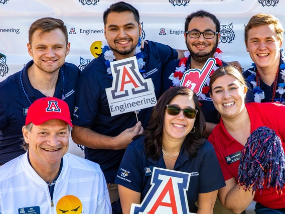 A group of seven people pose for a photo in front of a UA Engineering backdrop. They are smiling and holding props like a Block A and a foam finger in UA colors.