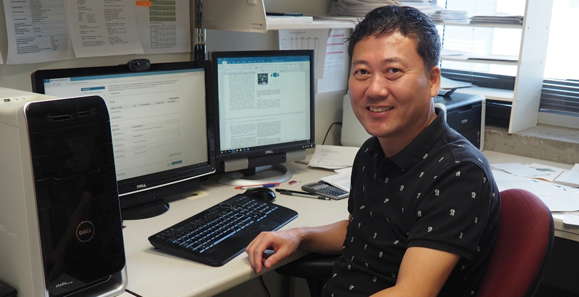 A man in a black polo sits at a desk in front of two computers.