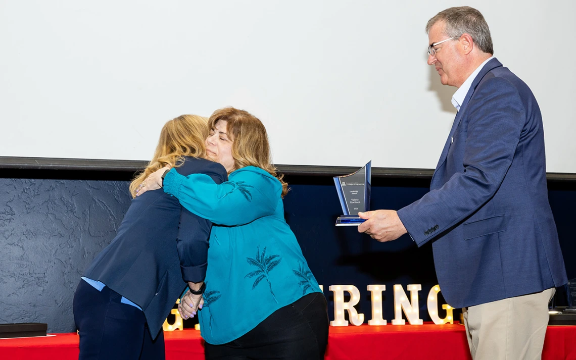 Two women in blue shirts embrace for a hug and one man in a blue blazer holds an award.