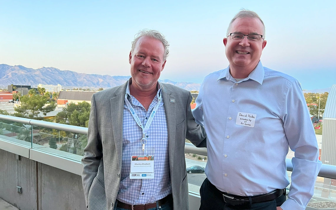 Two men with gray hair and blue dress shirts pose for a photo in front of a mountain range.