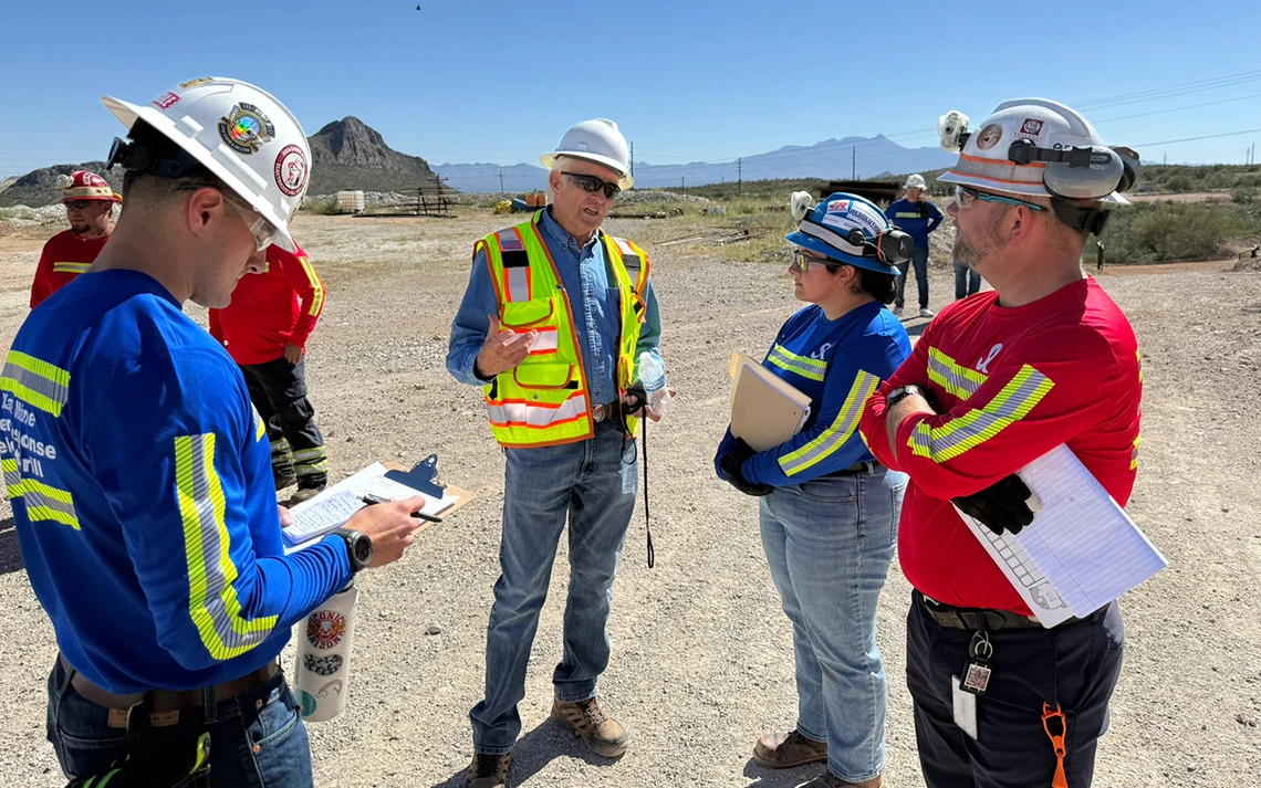 Five people in yellow vests stand on dusty land.