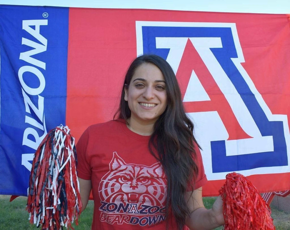 Maira Garcia smiles and holds two pom poms. Behind her is the University of Arizona Block A logo.