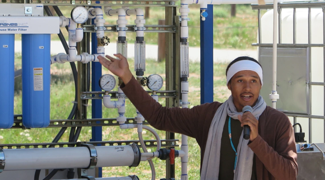 A young man holding a microphone gestures toward a systems of pipes and gauges.