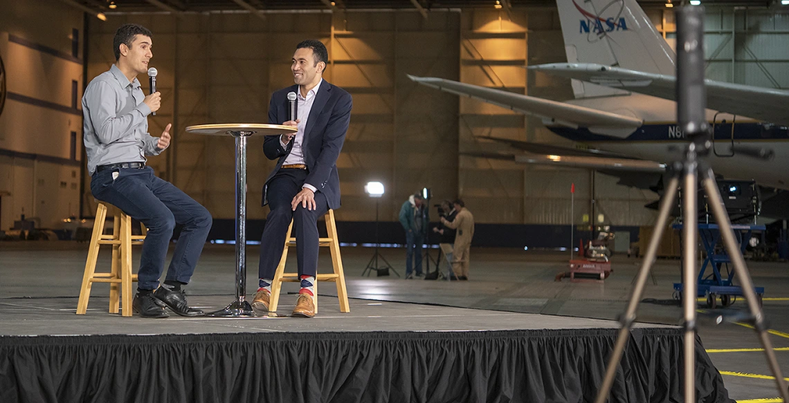 Two men sit on a stage with a NASA plane in the background