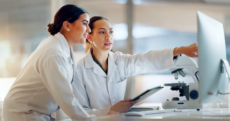 Two women in lab coats point at a computer