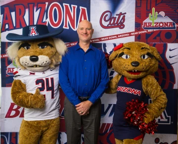 Villard and two mascots pose in front of a University of Arizona photo backdrop.