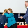 Two women in blue shirts embrace for a hug and one man in a blue blazer holds an award.
